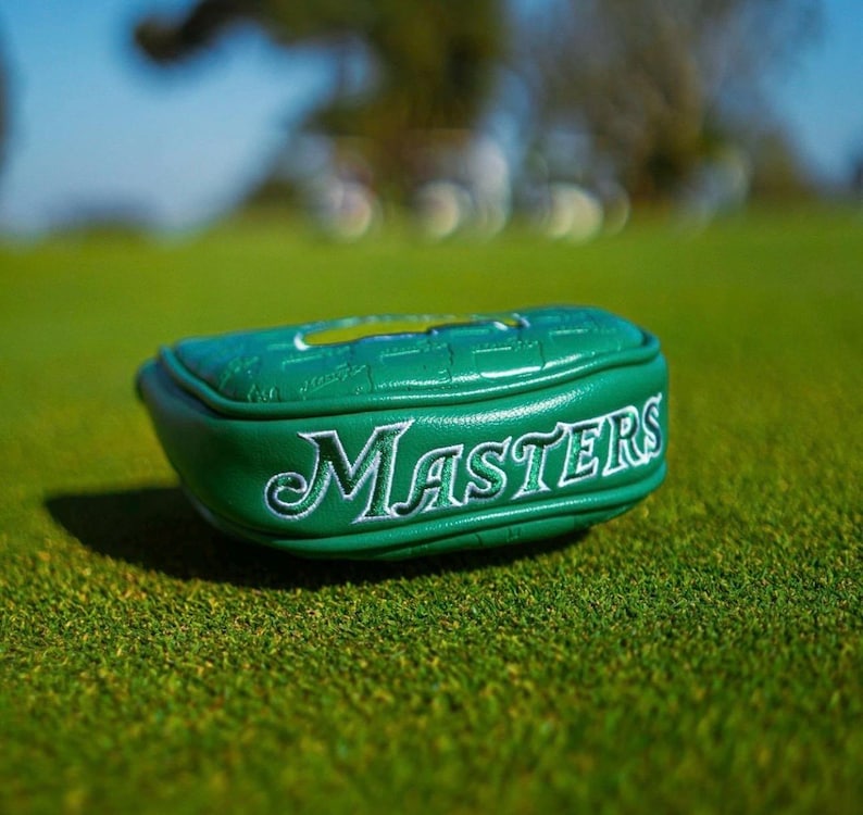 May include: A green golf club head cover with the word "Masters" embroidered in white. The cover is on a green golf course. The cover is made of a textured material.
