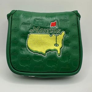 May include: Green golf putter cover with the Masters logo embroidered on the front. The logo features a yellow outline of the Augusta National Golf Club course, the word "Masters", and a red flag. The cover has a repeating embossed pattern of the word "Masters".