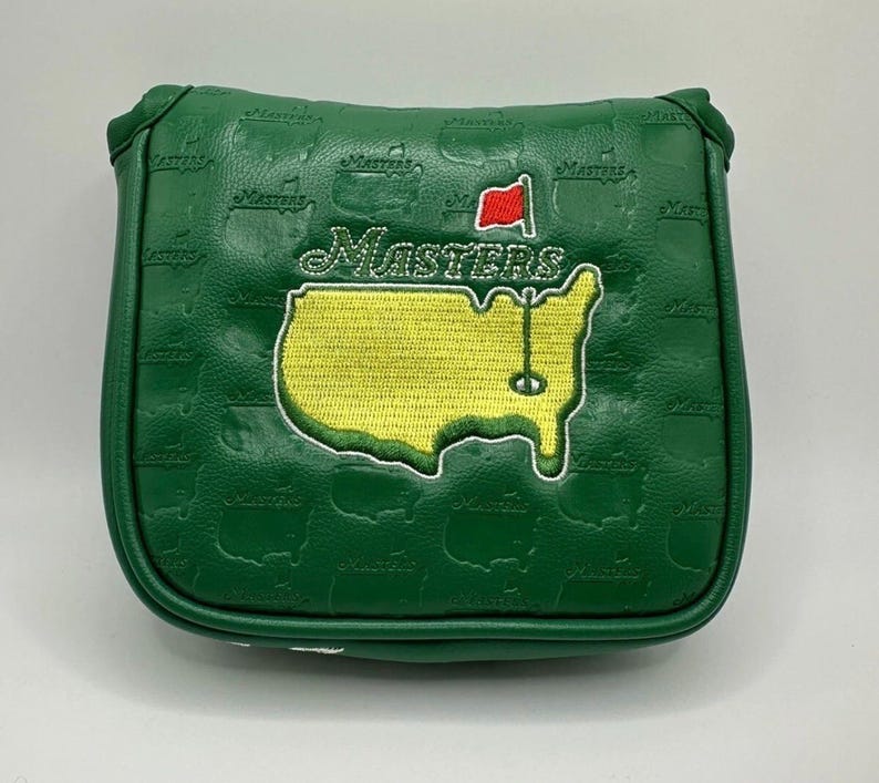 May include: Green golf putter cover with the Masters logo embroidered on the front. The logo features a yellow outline of the Augusta National Golf Club course, the word "Masters", and a red flag. The cover has a repeating embossed pattern of the word "Masters".