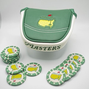 Puede incluir: Una funda para putter de golf verde y blanca y marcadores de bolas de golf. La funda del putter tiene la palabra "Masters" impresa, junto con un mapa amarillo del campo de golf. Los marcadores de bolas de golf tienen el mismo diseño.