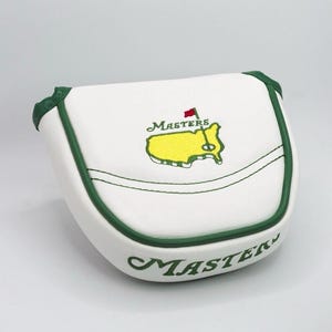 May include: White golf putter cover with green trim and the word "Masters" in green script. The cover features the Masters Tournament logo, a yellow outline of the Augusta National Golf Club with a red flag.