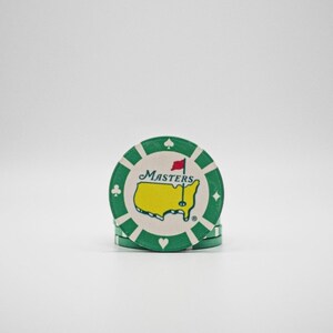 May include: A stack of green and white poker chips. Each chip features the Masters logo, a yellow outline of the United States with a red flag, and the word "Masters". The chips have suit symbols around the edge.