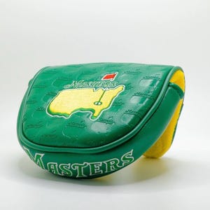 May include: A green golf putter cover with the word "Masters" in white lettering. The cover features a yellow and green map design with a red flag. The interior is yellow.