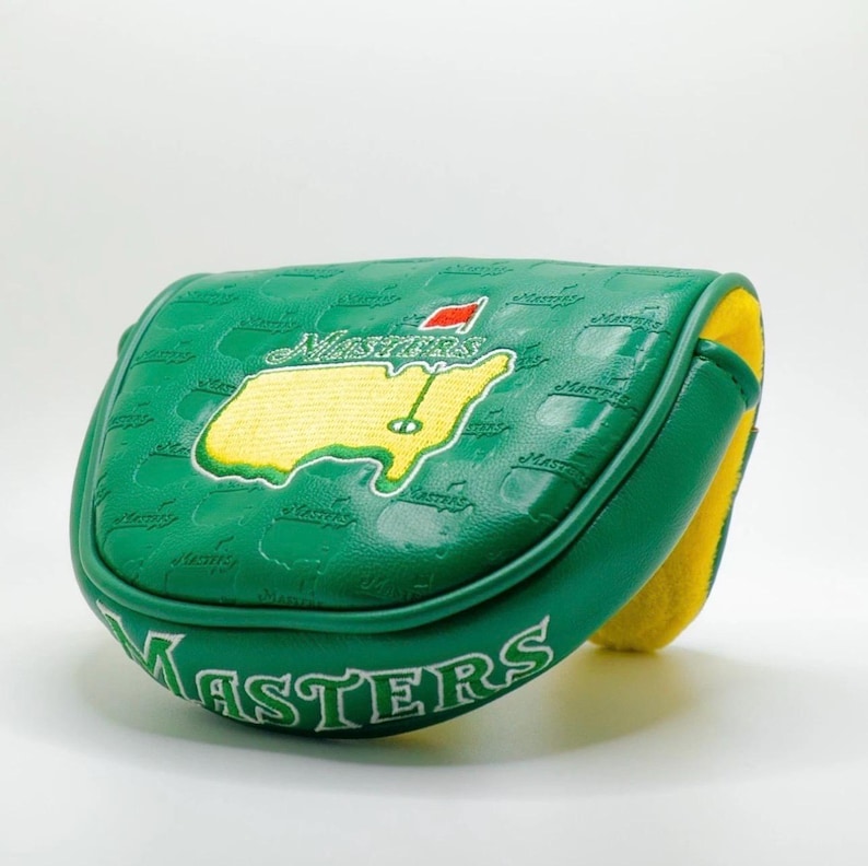 May include: A green golf putter cover with the word "Masters" in white lettering. The cover features a yellow and green map design with a red flag. The interior is yellow.