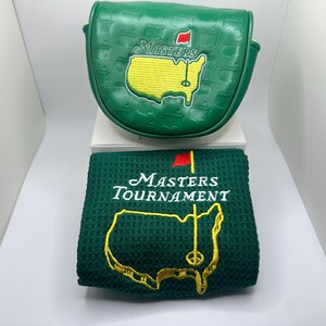 May include: A green golf club head cover and a matching green towel. Both items feature the Masters Tournament logo, including a yellow map of the course, a red flag, and the word "Masters" in white. The head cover has a textured pattern.
