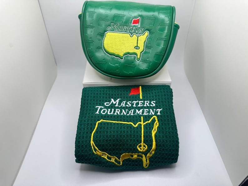 May include: A green golf club head cover and a matching green towel. Both items feature the Masters Tournament logo, including a yellow map of the course, a red flag, and the word "Masters" in white. The head cover has a textured pattern.