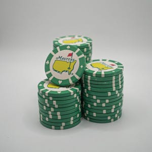 May include: Stacks of green and white poker chips with the Masters logo. The chips have a green border with white accents and the Masters logo in the center. The logo features a yellow outline of a golf course and the word "Masters".