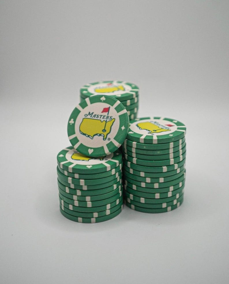 May include: Stacks of green and white poker chips with the Masters logo. The chips have a green border with white accents and the Masters logo in the center. The logo features a yellow outline of a golf course and the word "Masters".