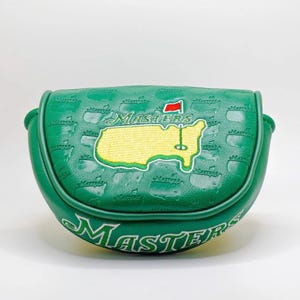May include: Green golf putter cover with the word "Masters" in white script. The cover features an embroidered yellow map of the Augusta National Golf Club with a red flag and a golf pin. The cover is textured with the word "Masters" repeated.