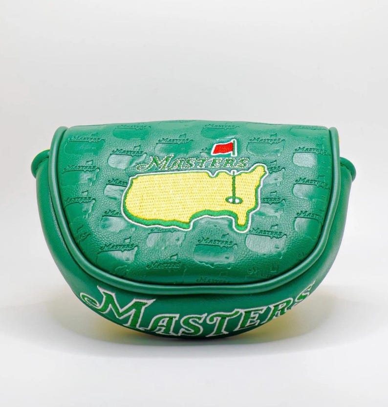 May include: Green golf putter cover with the word "Masters" in white script. The cover features an embroidered yellow map of the Augusta National Golf Club with a red flag and a golf pin. The cover is textured with the word "Masters" repeated.