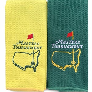 Può includere: Due asciugamani da golf, uno giallo e uno verde, con il logo del Masters Tournament ricamato. Il logo include le parole "Masters Tournament" sopra una mappa degli Stati Uniti con un segnaposto e una bandiera da golf.