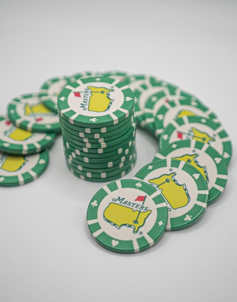 May include: A stack of green and white poker chips with the Masters logo. The chips feature a yellow outline of the state of Georgia with a red flag and the word "Masters". The chips are arranged in a semi-circle.