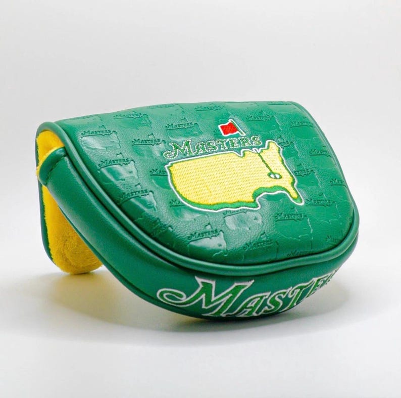 Puede incluir: Funda de putter de golf verde con la palabra "Masters" en escritura blanca. La funda presenta un mapa bordado en amarillo del campo de golf Augusta National y una bandera roja. La funda es verde con un interior amarillo.