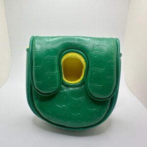 May include: A green golf putter cover with a textured pattern and yellow interior. The cover has a flap closure and a circular opening. The word "MASTERS" is embossed on the cover.