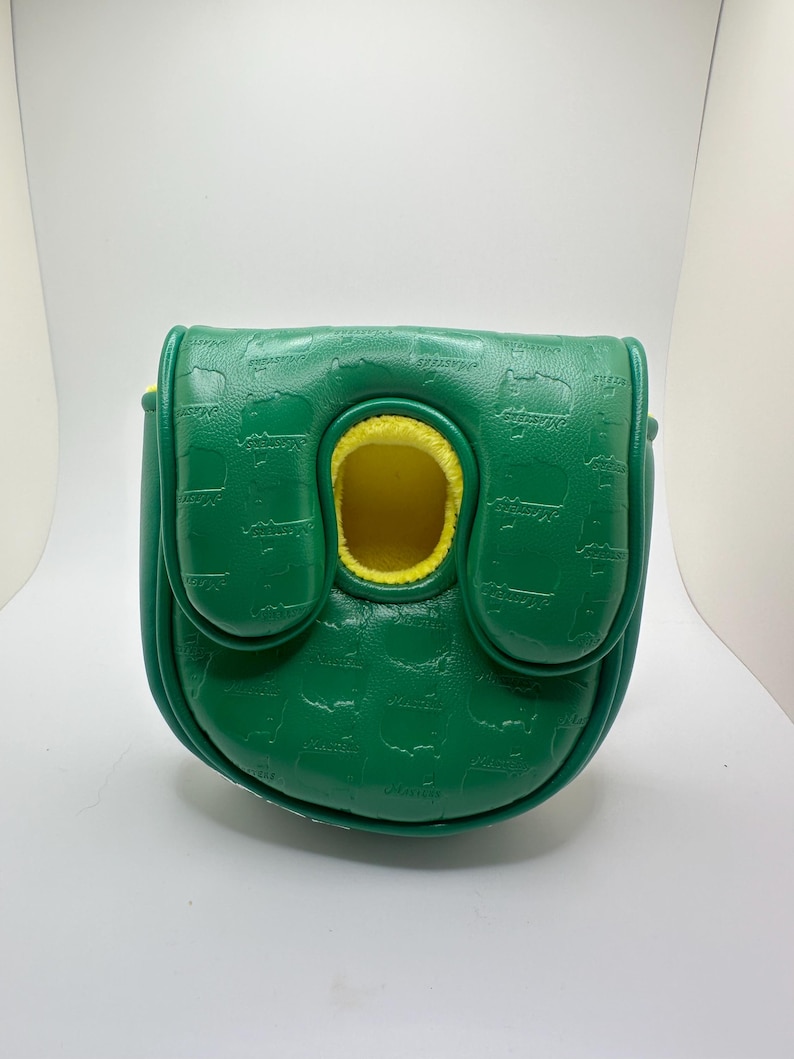 May include: A green golf putter cover with a textured pattern and yellow interior. The cover has a flap closure and a circular opening. The word "MASTERS" is embossed on the cover.