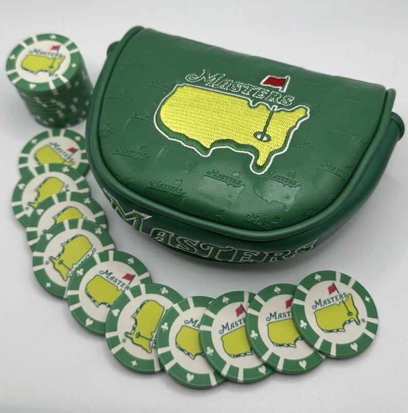 May include: A green golf putter cover and golf ball markers. The putter cover has the word "Masters" embroidered on it, along with a yellow and green map design. The golf ball markers have the same design.