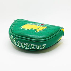 May include: Green golf putter cover with the word "MASTERS" in white lettering. The cover features a yellow and green embroidered map design. The cover is green with a textured pattern and yellow accents.