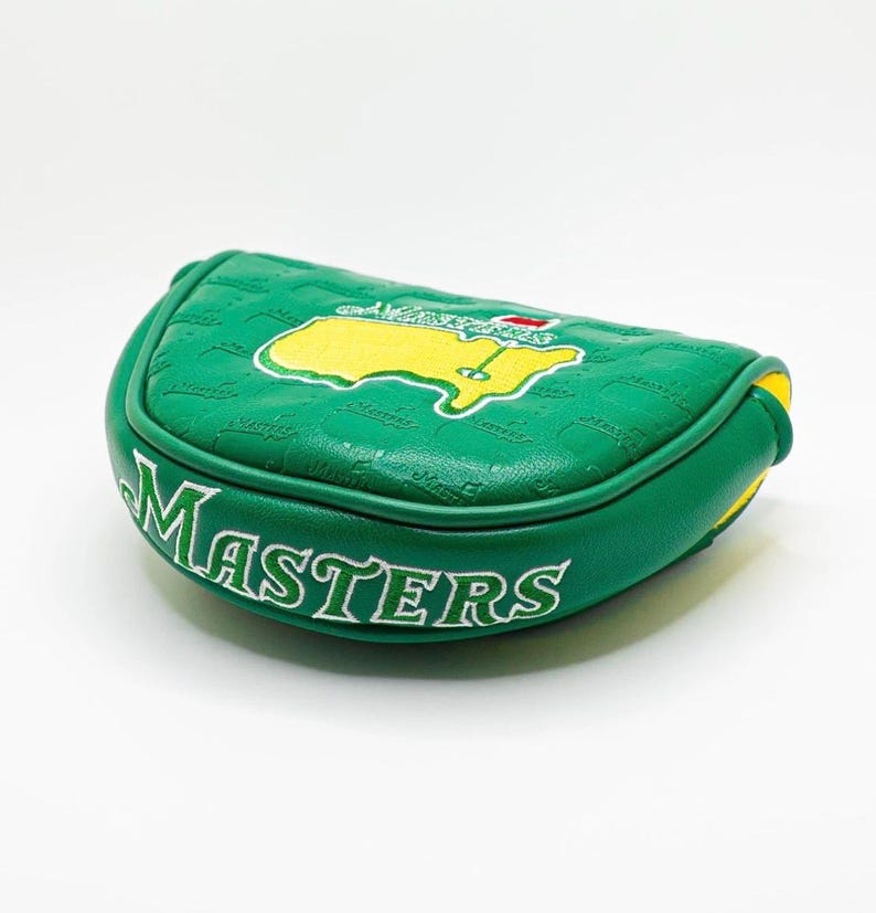 May include: Green golf putter cover with the word "MASTERS" in white lettering. The cover features a yellow and green embroidered map design. The cover is made of a textured material and has yellow trim.