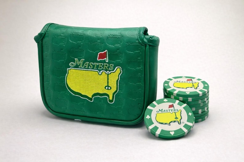 May include: Green golf putter cover and golf chips. The putter cover has the Masters logo in yellow and white. The golf chips are green and white with the Masters logo and a red flag.