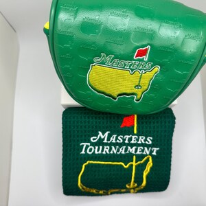 May include: Green golf club head cover and towel set. The head cover features the word "Masters" and a yellow outline of a map with a red flag. The towel has the words "Masters Tournament" and a yellow map outline.