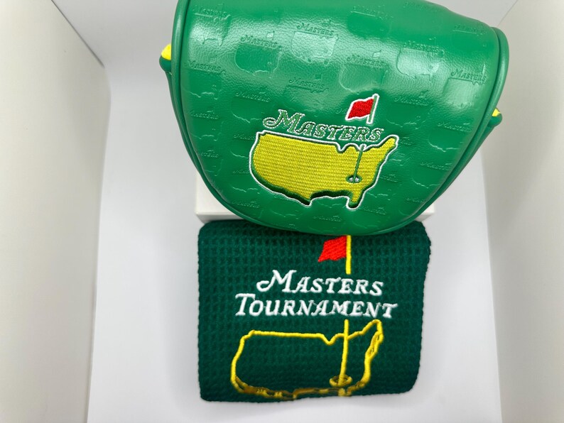 May include: Green golf club head cover and towel set. The head cover features the word "Masters" and a yellow outline of a map with a red flag. The towel has the words "Masters Tournament" and a yellow map outline.
