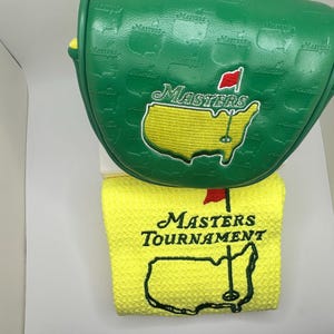 May include: Green golf club head cover and yellow golf towel. The head cover has the Masters logo embroidered in yellow and red. The towel has the Masters Tournament logo embroidered in black and red.