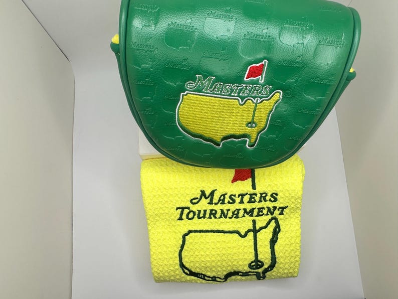May include: Green golf club head cover and yellow golf towel. The head cover has the Masters logo embroidered in yellow and red. The towel has the Masters Tournament logo embroidered in black and red.