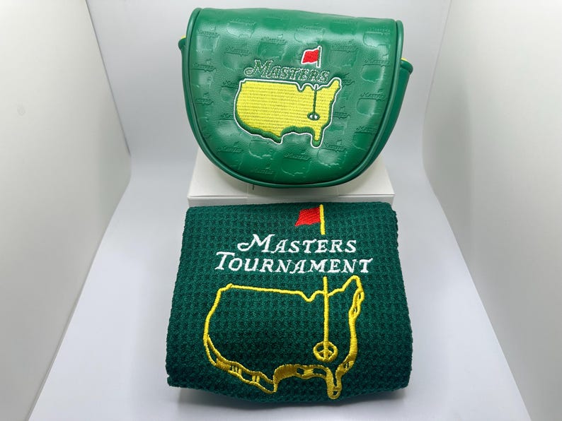 May include: A green golf club head cover and a matching green towel. Both items feature the Masters Tournament logo, including a yellow map of the United States with a red flag and the word "Masters" in white.