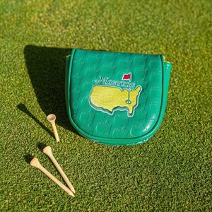 May include: A green golf club head cover with the Masters logo, featuring a yellow map of the course and the word "Masters". Three wooden golf tees are scattered on the green grass.