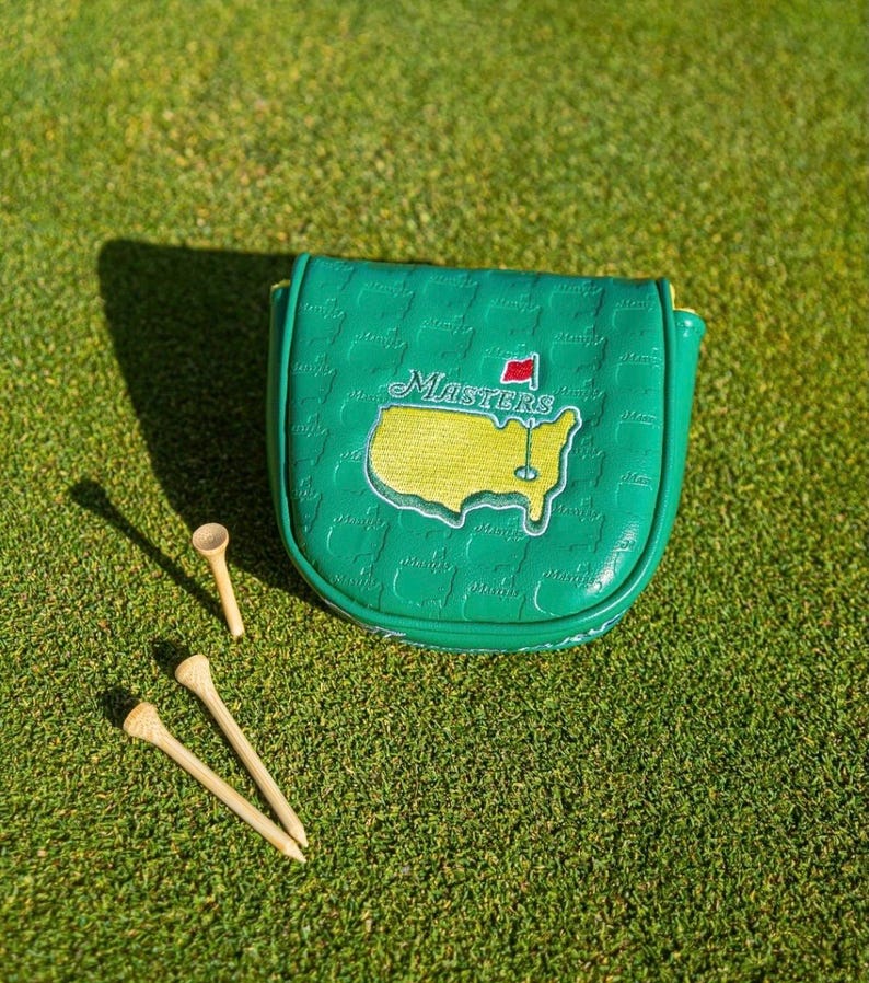 May include: A green golf club head cover with the Masters logo, featuring a yellow map of the course and the word "Masters". Three wooden golf tees are scattered on the green grass.