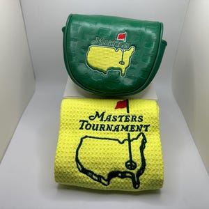 May include: A green golf club head cover and a yellow golf towel. The head cover features the Masters logo with a red flag and the outline of the United States. The towel has the Masters Tournament logo.