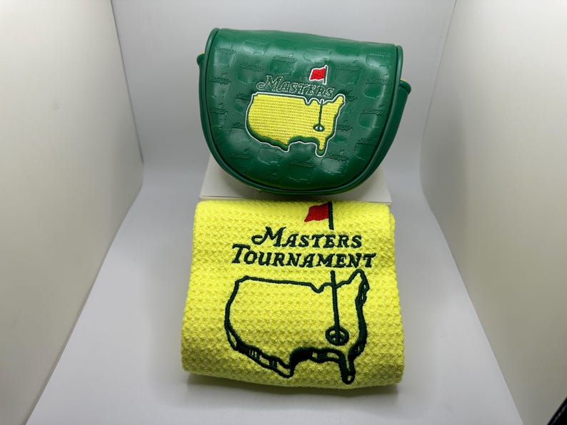 May include: A green golf club head cover and a yellow golf towel. The head cover features the Masters logo with a red flag and the outline of the United States. The towel has the Masters Tournament logo.