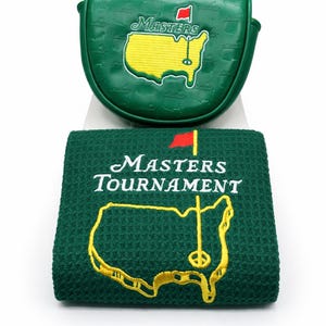 May include: Green golf putter cover and towel set. The putter cover and towel feature the Masters Tournament logo, including the outline of a golf course with a flag. The towel has the words "Masters Tournament" embroidered in white.