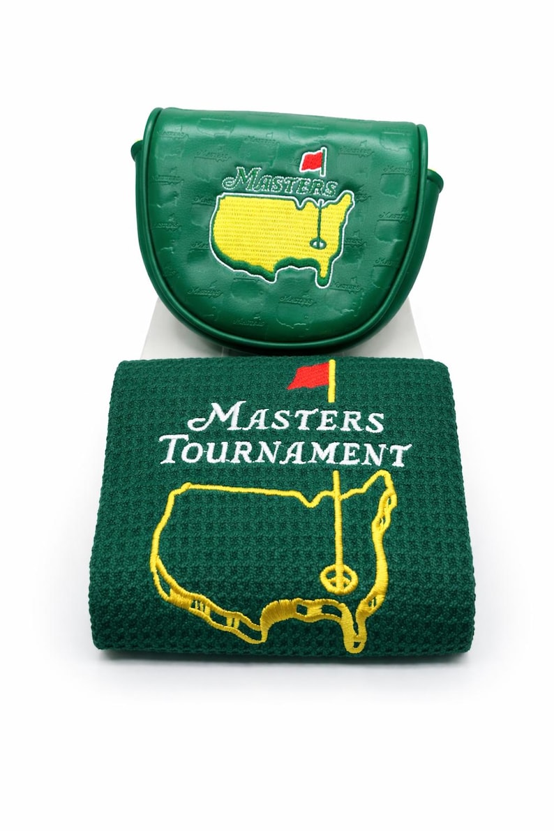 May include: Green golf putter cover and towel set. The putter cover and towel feature the Masters Tournament logo, including the outline of a golf course with a flag. The towel has the words "Masters Tournament" embroidered in white.