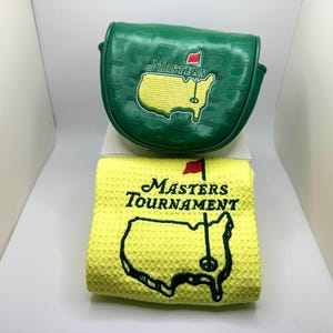 May include: A green golf club head cover and a yellow golf towel. The head cover has the word "MASTERS" and a map of the golf course embroidered on it. The towel has the words "MASTERS TOURNAMENT" and a map of the golf course.