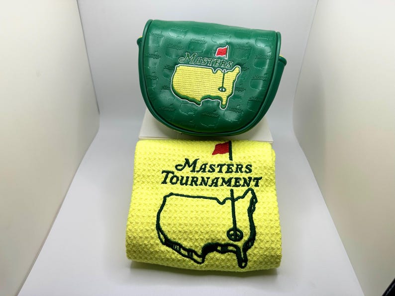 May include: A green golf club head cover and a yellow golf towel. The head cover has the word "MASTERS" and a map of the golf course embroidered on it. The towel has the words "MASTERS TOURNAMENT" and a map of the golf course.