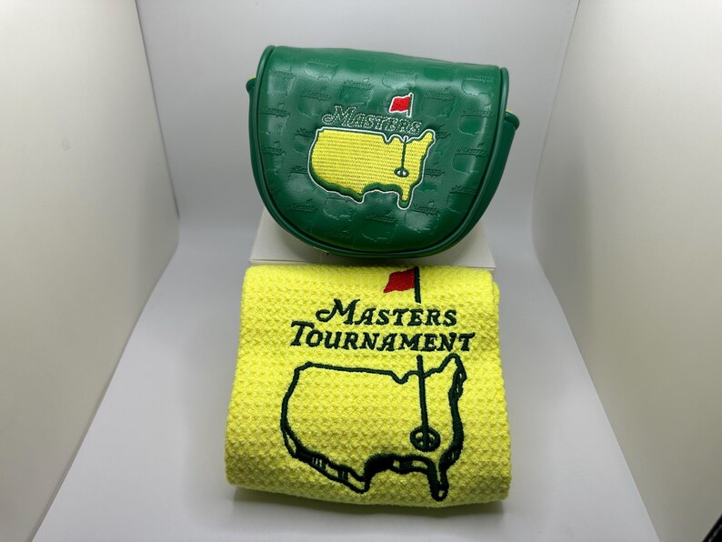 May include: Green golf club cover and yellow golf towel. The cover has the word "Masters" and a map of Georgia with a red flag. The towel has the words "Masters Tournament" and a map of Georgia with a golf pin.