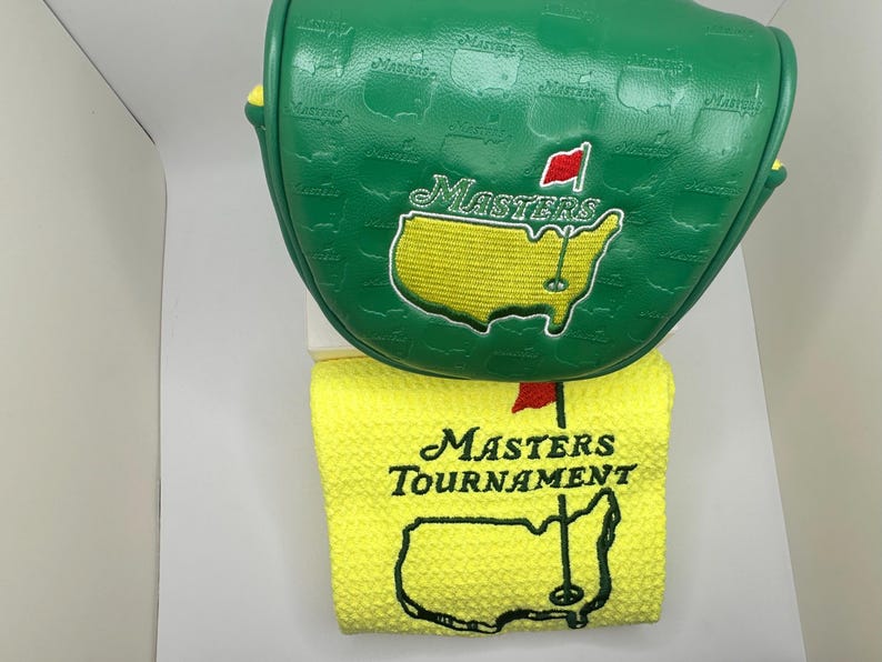 May include: Green golf club head cover and yellow golf towel. The head cover has the word "Masters" and a map of the course embroidered on it. The towel has the words "Masters Tournament" and a map of the course embroidered on it.