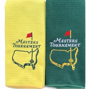 Puede incluir: Dos toallas de golf, una amarilla y otra verde, con el logotipo del Masters Tournament bordado. El logotipo incluye las palabras "Masters Tournament", una bandera roja y un mapa de los Estados Unidos con un pin de golf. Las toallas tienen un tejido de gofre texturizado.