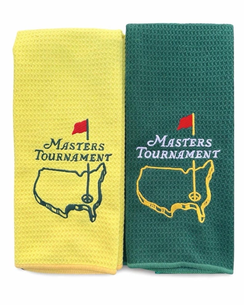 Puede incluir: Dos toallas de golf, una amarilla y otra verde, con el logotipo del Masters Tournament bordado. El logotipo incluye las palabras "Masters Tournament", una bandera roja y un mapa de los Estados Unidos con un pin de golf. Las toallas tienen un tejido de gofre texturizado.