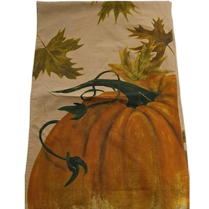 May include: A rectangular table runner featuring a beige background with a large orange pumpkin illustration. The design includes dark green stems and curling vines, accented by scattered brown and green maple leaves across the fabric surface.