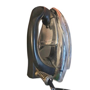 May include: Vintage General Electric iron with a black handle and a chrome-plated base. The iron has a spring-loaded cord protector and the General Electric logo is visible on the front.