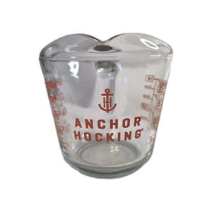 Könnte beinhalten: Ein Messbecher aus klarem Glas mit dem orangefarbenen Anchor Hocking-Logo und einem Anker-Symbol auf der Vorderseite. Die Außenfläche zeigt abgestufte Messmarkierungen in Unzen und Millilitern sowie einen breiten Ausguss an der Oberseite.