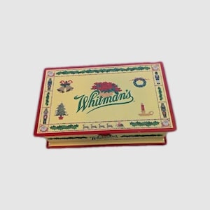 Könnte beinhalten: Eine rechteckige Whitman's Süßwarenschachtel mit cremefarbenem Deckel, verziert mit einem grünen Schriftzug-Logo, einem roten Weihnachtsstern und weihnachtlichen Motiven wie Nussknackern, Glocken, einem Tannenbaum, einer Kerze und Rentieren.