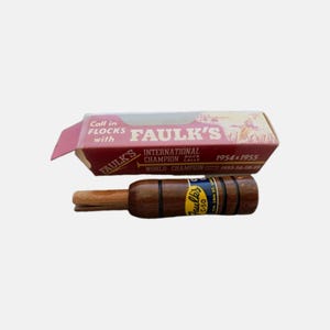 以下が含まれることがあります： ダークブラウン仕上げで黒いリングが付いた木製のダックコール。Faulk's C-50と記された青と黄色のラベルが貼られています。Faulk's International Champion Duck Callsと印刷された長方形の段ボール箱が付属しています。