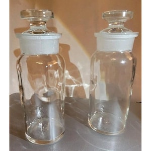 Puede incluir: Dos frascos de boticario de vidrio transparente con tapones de vidrio. Los frascos tienen forma cilíndrica con una base más ancha y un cuello más estrecho. Los tapones son de vidrio transparente a juego con una parte superior plana y un borde blanco.