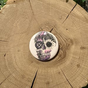 May include: A round, off-white decorative stone featuring a detailed skull design. The skull is adorned with floral elements, a sunflower, and a checkered pattern, with a color palette of black, pink, and purple.