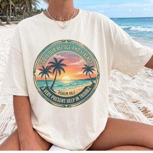 Op de afbeelding: Een crèmekleurig T-shirt met korte mouwen en een ronde afbeelding van een tropische zonsondergang op het strand met palmbomen. De tekst luidt: GOD IS OUR REFUGE AND STRENGTH A VERY PRESENT HELP IN TROUBLE PSALM 46:1 in oranje, blauwe en groene tinten.