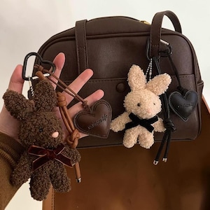 Op de afbeelding: Drie pluche konijnensleutelhangers in bruin en beige, elk met een hartvormig label. Het bruine konijn heeft een rode strik, terwijl het beige konijn een zwarte strik heeft. De labels lezen "xallan-life". Een bruine handtas staat op de achtergrond.