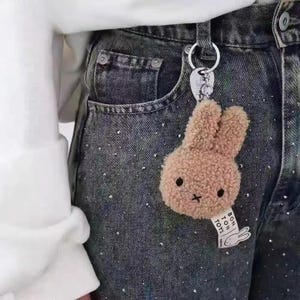 以下が含まれることがあります： 濃い色のデニムジーンズに付いた、薄茶色のウサギのぬいぐるみキーホルダー。ウサギは小さな黒い十字の口と黒い目があります。「BON TON TOYS」と書かれたタグがキーホルダーに付いています。